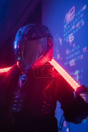 Foto de Una figura femenina, vestida con un conjunto negro táctico futurista y un casco de motocicleta, empuña dos lámparas de neón mientras se coloca delante de una pantalla de símbolos digitales proyectados - Imagen libre de derechos