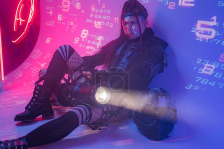 Téléchargez les photos : Une femme en costume noir tactique futuriste repose sur le sol, avec un casque de moto et un projecteur massif à côté d'elle, sur fond de symboles numériques projetés - en image libre de droit