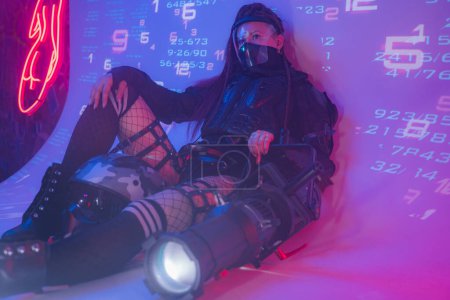 Téléchargez les photos : Une femme en costume noir tactique futuriste repose sur le sol, avec un projecteur massif à côté d'elle, sur fond de symboles numériques projetés, avec un espace de copie - en image libre de droit