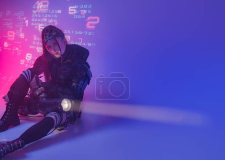Foto de Una figura femenina, vestida con un conjunto negro táctico futurista, se reclina en el suelo con un proyector considerable sobre un fondo de símbolos digitales proyectados - Imagen libre de derechos