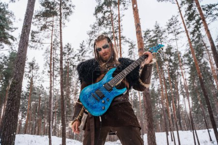 Téléchargez les photos : Un musicien d'inspiration viking joue de la guitare électrique dans une forêt enneigée, incarnant une fusion entre l'ancien guerrier et la culture musicale moderne. - en image libre de droit