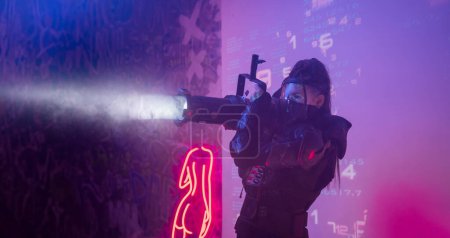 Foto de Una mujer con un traje negro táctico futurista sostiene un gran proyector que se asemeja a un cañón de luz, de pie sobre un telón de fondo de símbolos digitales proyectados - Imagen libre de derechos