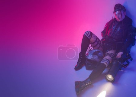 Foto de Una mujer con un traje negro táctico futurista yace en el suelo, con un proyector masivo a su lado, sobre un telón de fondo de símbolos digitales proyectados, con espacio para copiar - Imagen libre de derechos
