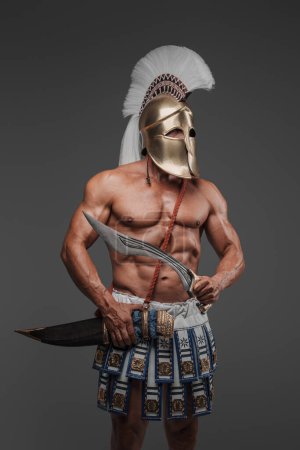 Téléchargez les photos : Portrait d'un ancien grec hoplite, posant dans un studio avec un torse musculaire nu, portant un casque, et tenant un gladius, sur un fond gris - en image libre de droit