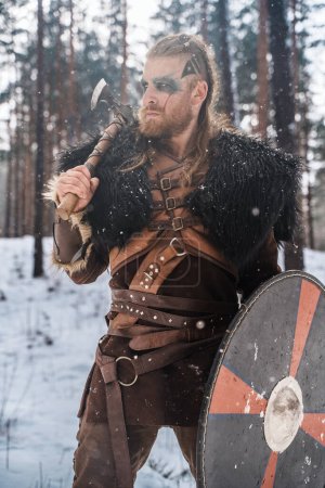 Téléchargez les photos : Un guerrier viking féroce, armé d'une hache et d'un bouclier dans une pinède enneigée, représentant la force et la culture nordique historique - en image libre de droit