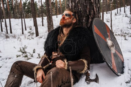 Téléchargez les photos : Un guerrier viking avec de la peinture de guerre sur le visage repose contre un arbre dans la neige, paraissant blessé et épuisé - en image libre de droit