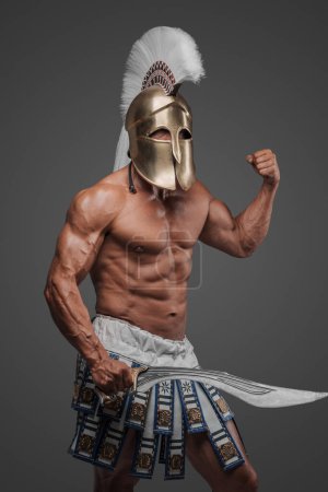 Photo for Portrait of an ancient Greek hoplite, posing in a studio with a bare muscular torso, wearing a helmet, and holding a gladius, set against a gray background - Royalty Free Image