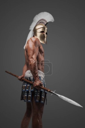 Photo for An impressive portrait of a bare-chested ancient Greek hoplite, wearing a helmet and brandishing a spear, standing in a studio with a gray backdrop - Royalty Free Image