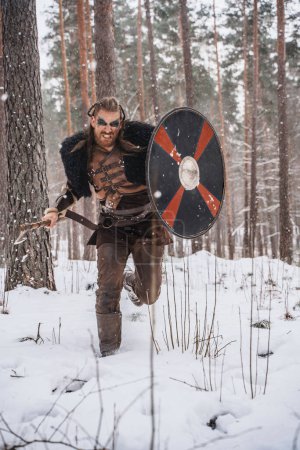 Foto de Imagen dinámica de un guerrero vikingo cargando con hacha y escudo en medio de la nieve cayendo en un denso bosque - Imagen libre de derechos