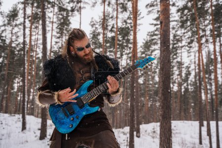 Téléchargez les photos : Un musicien d'inspiration viking joue de la guitare électrique dans une forêt enneigée, incarnant une fusion entre l'ancien guerrier et la culture musicale moderne. - en image libre de droit