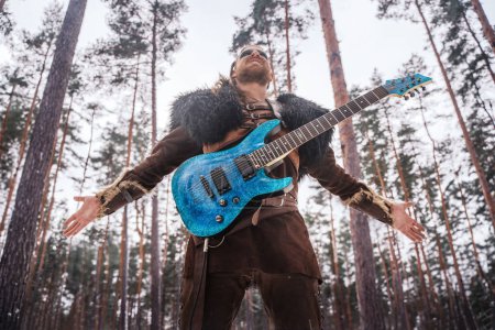 Foto de Un músico de inspiración vikinga toca una guitarra eléctrica en un bosque nevado, encarnando una fusión de guerrero antiguo y cultura musical moderna - Imagen libre de derechos