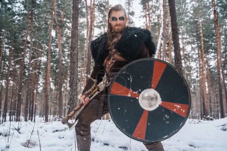 Téléchargez les photos : Un homme habillé en guerrier viking se tient debout avec une hache et un bouclier dans une forêt enneigée, évoquant l'imagerie nordique historique - en image libre de droit