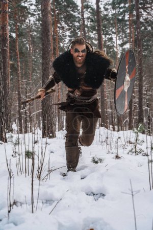 Téléchargez les photos : Image dynamique d'un guerrier viking chargé de hache et de bouclier au milieu d'une neige tombante dans une forêt dense - en image libre de droit
