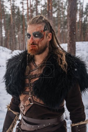 Téléchargez les photos : Gros plan d'un guerrier viking avec une peinture de guerre frappante, debout dans une forêt enneigée, exsudant une présence féroce et couvante - en image libre de droit