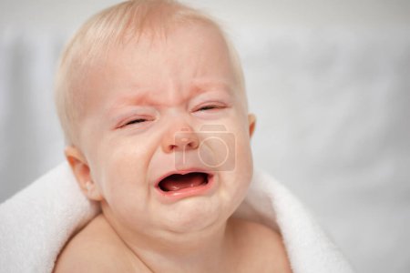 Téléchargez les photos : Bébé pleurant mignon avec les yeux fermés. Petit garçon triste - portrait rapproché. Enfant criant couvert de serviette de bain. - en image libre de droit