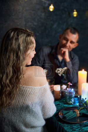 Photo for A beautiful couple sits at a candlelit table in a dark restaurant. The man gazes thoughtfully at his partner while she sits in the foreground, surrounded by flickering candlelight. - Royalty Free Image