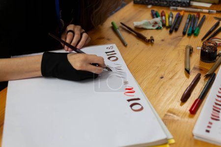Erfahrene Kalligraphin übt sich in Schreiben und Zeichnen von Schriften im Renaissance-Stil in einem Skizzenbuch. Auf dem Holztisch liegen Marker und Buntstifte