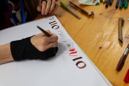 Experienced calligrapher woman practices writing and drawing Renaissance-style fonts in a sketchbook. On the wooden table, there are markers and colored pencils