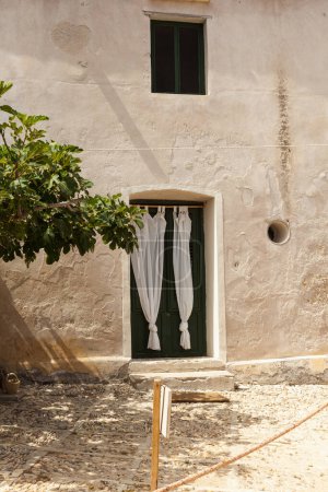View of an old house with white curtains on the door, next to a centuries-old fig tree, Tonnara di Scopello, Sicily.