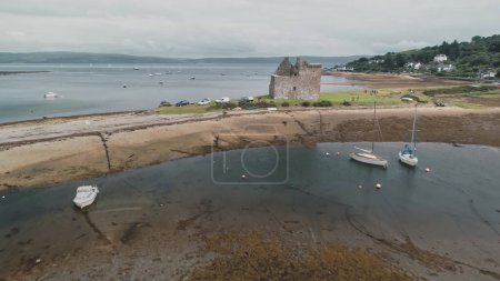 Sea gulls flying at brown sand beach on ocean shore aerial. Nature seascape with historical ancient castle ruins. Seagulls bird at summer cloudy day. Ships, yachts at harbor of Arran Island, Scotland