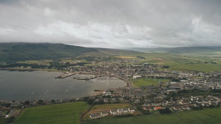 Port town at ocean bay aerial. Traffic highway at sea shore. Modern and historic buildings at pier city Campbeltown, Scotland, Europe. Boats, yachts, ships at dock. Green landscape at highland island
