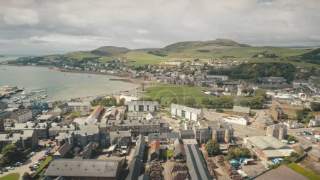 Aerial Campbeltown sun cityscape at green forest mountains. Modern buildings at urban downtown streets. Traffic roads with , trucks drives. European city scape at summer sunny day. Cinematic soft shot