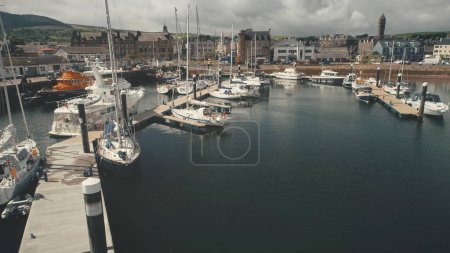 Closeup harbor dock with yachts aerial. Ships and sailboats at marina on ocean bay. Water transport at port city of Campbeltown cityscape. Scotland seascape at summer day. Cinematic drone shot