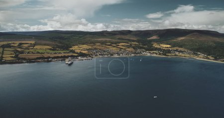 Vista aérea del muelle del transbordador marítimo de Brodick: barcos, barcos y veleros en la superficie del océano en el puerto. Maravilloso paisaje escocés de la naturaleza con bosques y valles en tiro de verano