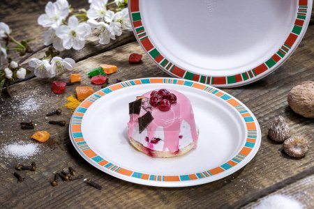 Foto de Tarta de bayas con crema rosa chocolate negro en un plato redondo ecológico desechable sobre un fondo de madera texturizada - Imagen libre de derechos