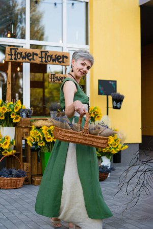 Foto de Elegante mujer adulta con una cesta de flores cerca de un café - una tienda de flores. Girasoles y lavanda. - Imagen libre de derechos