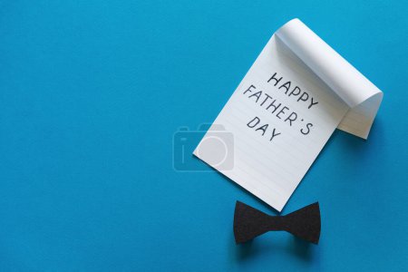 Foto de Feliz Concepto del Día de los Padres. Hoja de papel con felicitaciones. Negro elegante corbata de lazo de papel y cuaderno sobre fondo azul. Copiar espacio. - Imagen libre de derechos