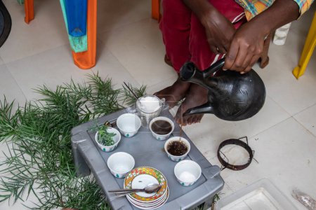 Foto de A jebena sits on a grass-covered table with small, handle-less cups arranged around it. Freshly roasted coffee beans, a mortar and pestle, and incense smoke create a traditional Ethiopian coffee ceremony setup. - Imagen libre de derechos