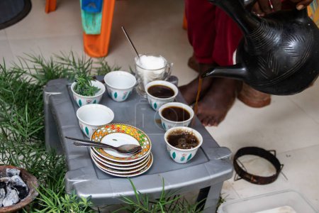 A jebena sits on a grass-covered table with small, handle-less cups arranged around it. Freshly roasted coffee beans, a mortar and pestle, and incense smoke create a traditional Ethiopian coffee ceremony setup.
