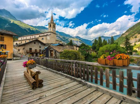 View of the wooden bridge over alpine river as church and houses on background in small surrounded by the mountains town of Val-Cenis, France.