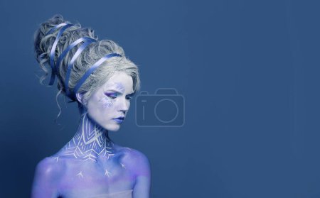 Gorgeous woman Snow Queen with creative makeup, perfect hairstyle and painting snowflake on her skin against blue studio wall background. Carnival or Halloween party makeup, studio portrait