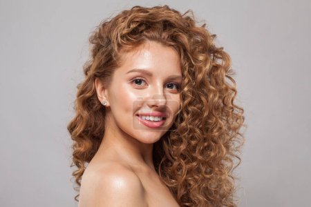 Fashion model with wavy hairstyle smiling. Attractive young woman with curly hair posing at studio. Face of a beautiful woman with long brown curly hair. Female face with natural makeup