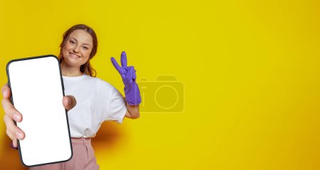 Photo for Attractive woman in cleaning gloves holding smartphone with empty white screen display for new Application and showing thumb up on bright yellow studio wall background. Mockup banner - Royalty Free Image
