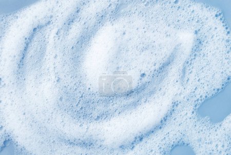 Téléchargez les photos : Bulles blanches de mousse nettoyante pour la peau sur fond bleu clair. Savon, shampooing ou gel douche texture mousse, gros plan, vue sur le dessus - en image libre de droit