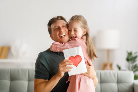 Foto de La hija del niño felicita a su padre y le da una postal. Papá y la niña sonríen y se abrazan. Vacaciones familiares y unión. - Imagen libre de derechos