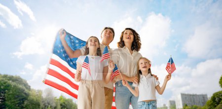 Foto de Vacaciones patrióticas. Familia feliz, padres e hijas niñas niñas con bandera americana al aire libre. Estados Unidos celebra el 4 de julio. - Imagen libre de derechos