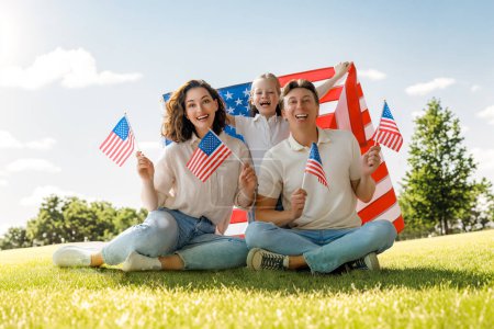Foto de Vacaciones patrióticas. Familia feliz, padres e hija niña con bandera americana al aire libre. Estados Unidos celebra el 4 de julio. - Imagen libre de derechos