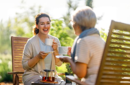 Photo for Happy young woman and her mother drinking tea in summer morning. Family sitting in the garden with cups and enjoying the conversation. - Royalty Free Image