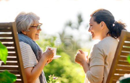 Téléchargez les photos : Joyeux jeune femme et sa mère buvant du thé le matin d'été. Famille assise dans le jardin avec des tasses et profiter de la conversation. - en image libre de droit