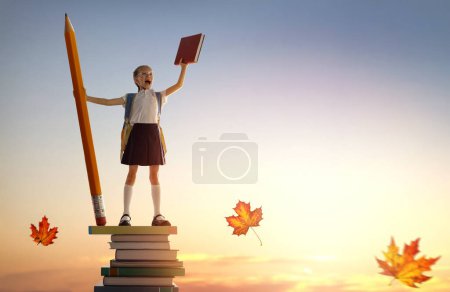 Téléchargez les photos : Retourne à l'école ! Joyeux enfant industrieux mignon debout sur la tour des livres et tenant un énorme crayon sur fond de ciel couchant. Concept d'éducation et de lecture. Le développement de l'imagination. - en image libre de droit
