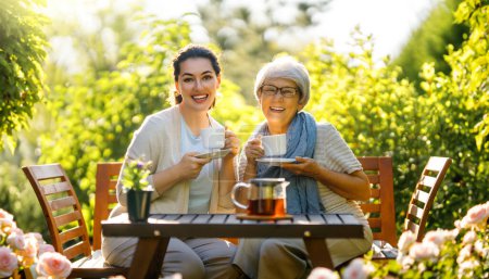 Téléchargez les photos : Joyeux jeune femme et sa mère buvant du thé le matin d'été. Famille assise dans le jardin avec des tasses et profiter de la conversation. - en image libre de droit