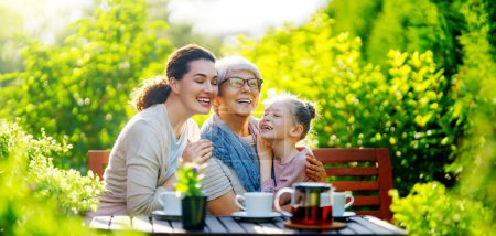 Foto de Feliz niña, su madre y su abuela tomando té en la mañana de verano. Familia sentada en el jardín con copas y disfrutando de la conversación. - Imagen libre de derechos