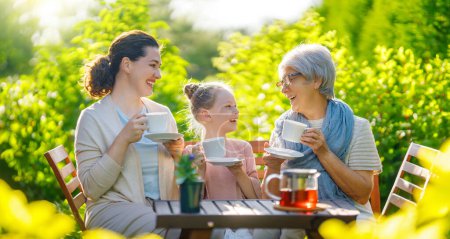 Foto de Feliz niña, su madre y su abuela tomando té en la mañana de verano. Familia sentada en el jardín con copas y disfrutando de la conversación. - Imagen libre de derechos
