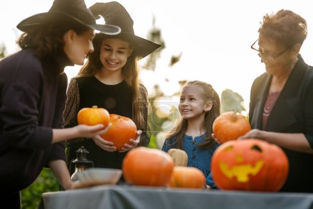 Téléchargez les photos : Bonne famille se préparant pour Halloween. Mère, grand-mère et enfants sculptant des citrouilles dans la cour arrière de la maison. - en image libre de droit