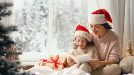 Téléchargez les photos : Joyeux Noël et Joyeuses Fêtes. Joyeux maman et sa fille mignonne fille lecture d'un livre. Parent et petit enfant s'amusent près de l'arbre à l'intérieur. Matin Noël. - en image libre de droit