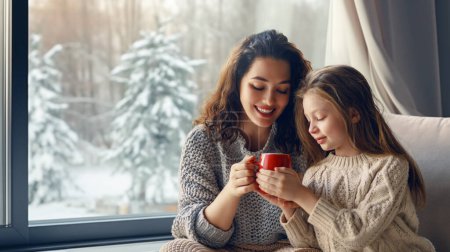 Photo for Happy loving family. Mother and daughter are hugging and drinking hot beverage enjoying winter nature in the  window. - Royalty Free Image